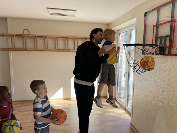 Ein Dragonspieler hebt ein Kindergartenkind mit Basketball hoch zum Korb. Zwei andere Kinder stehen und warten, dass sie dran sind.